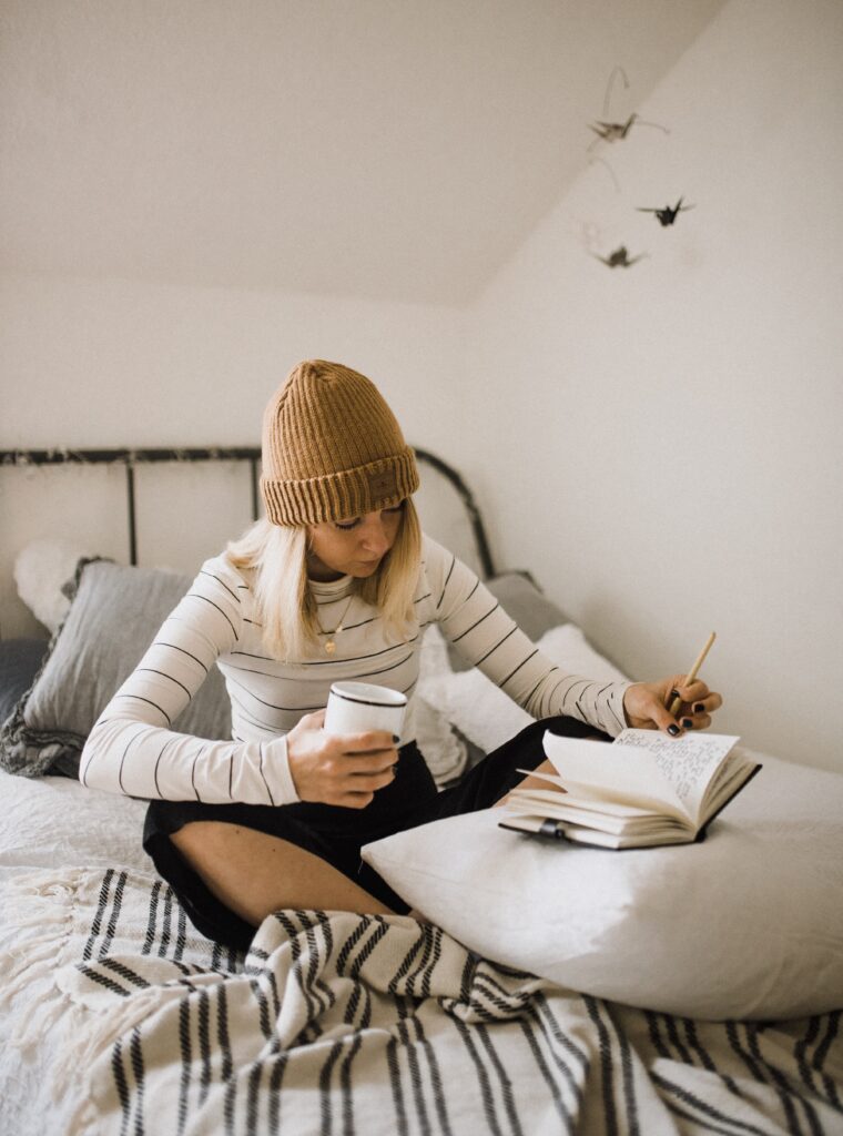 Vrouw zit op bed, meditatief en schrijvend, met een kop koffie in de hand. Schrijven moet je doen om helder en duidelijk schrijven te verbeteren.